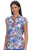 A woman modeling a short-sleeve collared scrub top with a colorful floral and butterfly print on a white background, paired with blue bottoms, against a light gray studio backdrop.