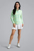 A woman modeling a long-sleeve quarter-zip golf top with a light green and yellow tie-dye pattern, paired with a white golf skirt and white golf shoes, against a solid grey studio background.