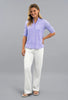 A woman modeling a light purple and white patterned short-sleeve quarter-zip top with white pants, standing against a solid  studio background.